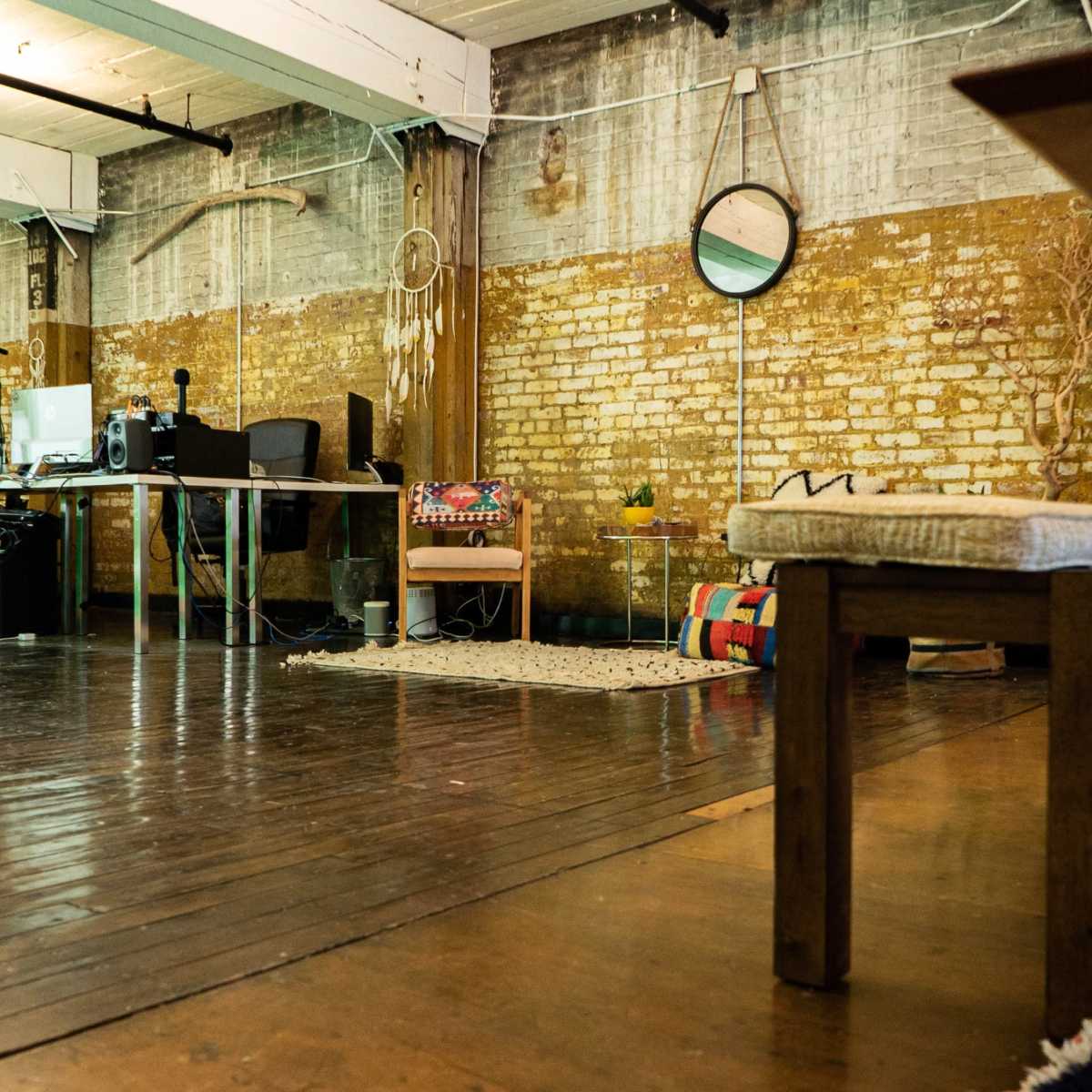 An industrial-style workspace with wooden flooring, exposed brick walls, and various furniture items, including a bench and a desk setup.