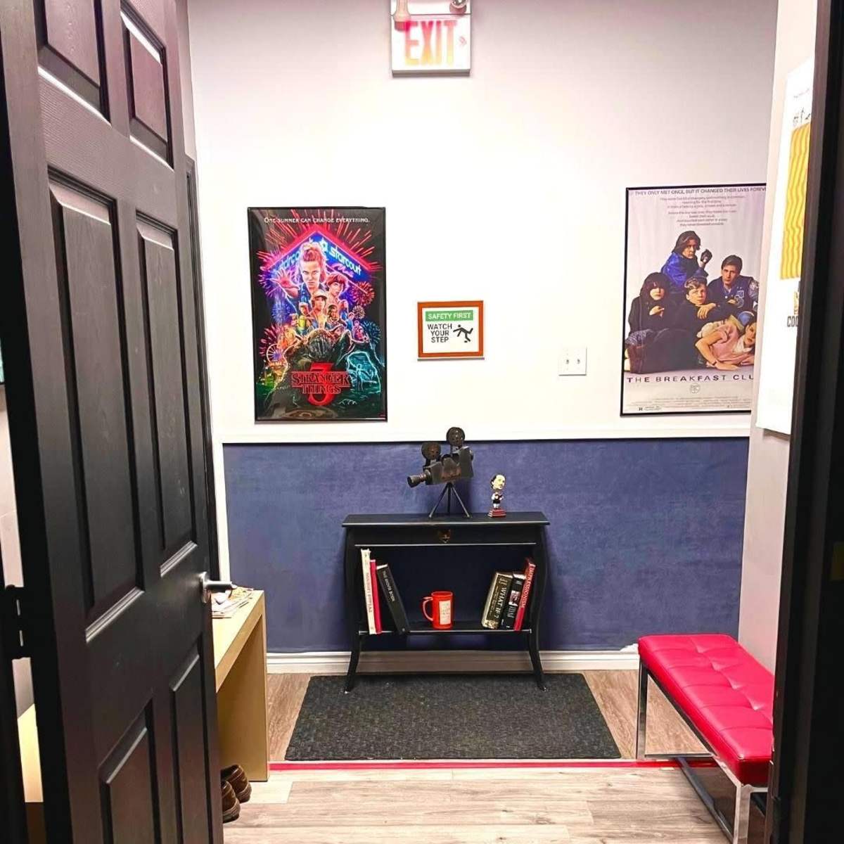An office entrance featuring a black door, a blue wall, a small bookshelf with a red mug, and two movie posters on the walls.