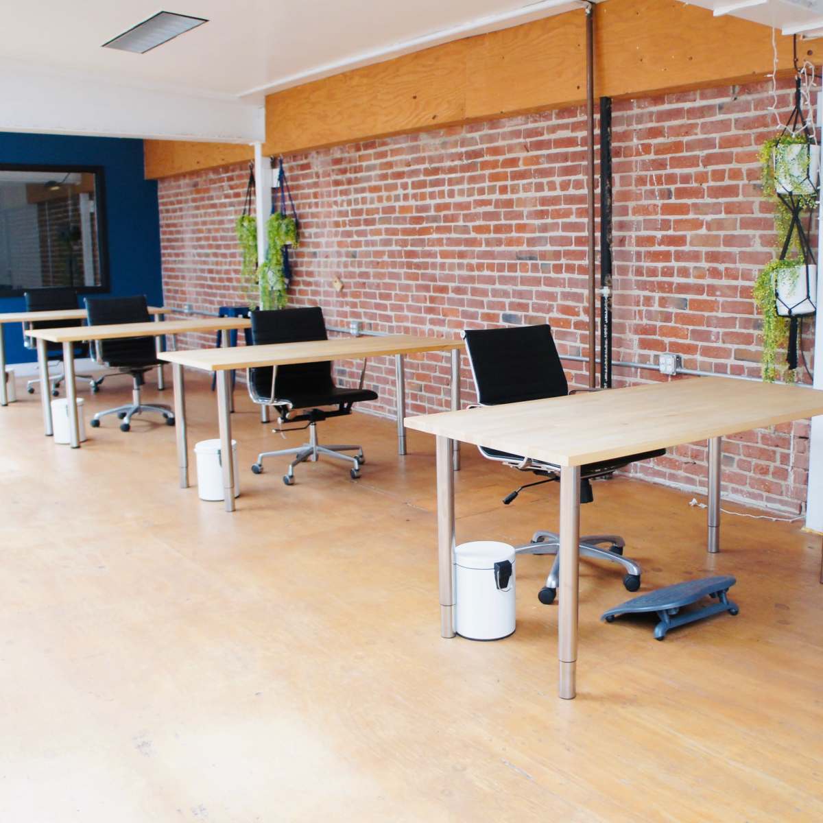 A workspace with several wooden desks and rolling chairs against a backdrop of exposed brick walls and hanging plants.
