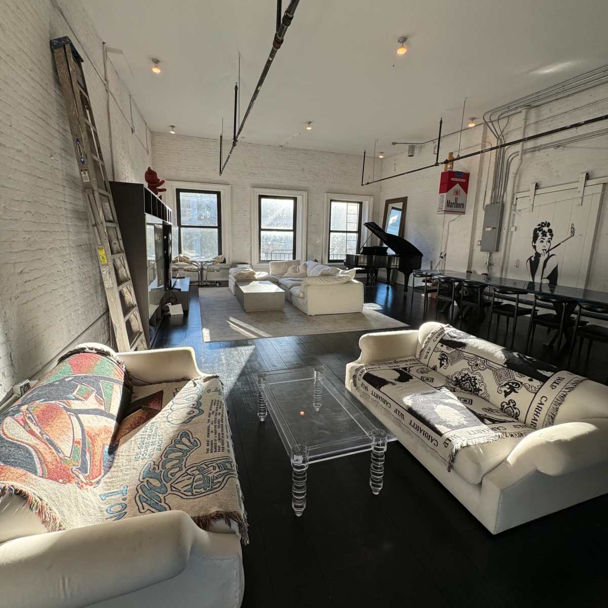 The room features several white couches covered with blankets, a glass coffee table, a piano, and large windows, all set against a backdrop of exposed brick walls and black flooring.