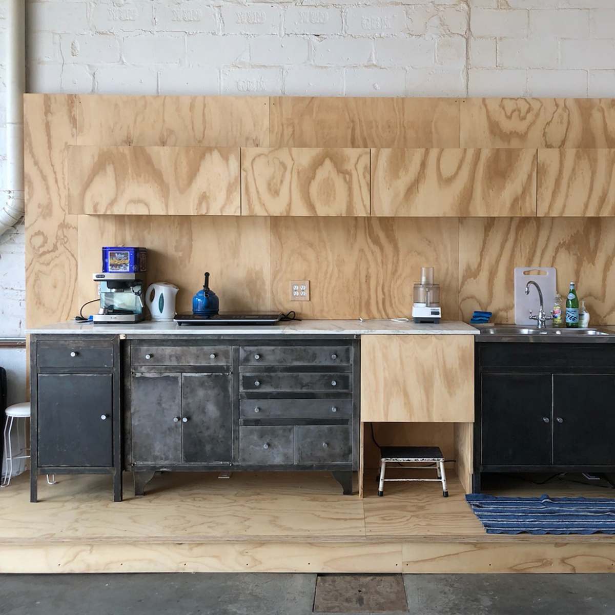 A minimalist kitchen with a wooden wall, metal cabinets, a sink, and a stainless steel refrigerator.