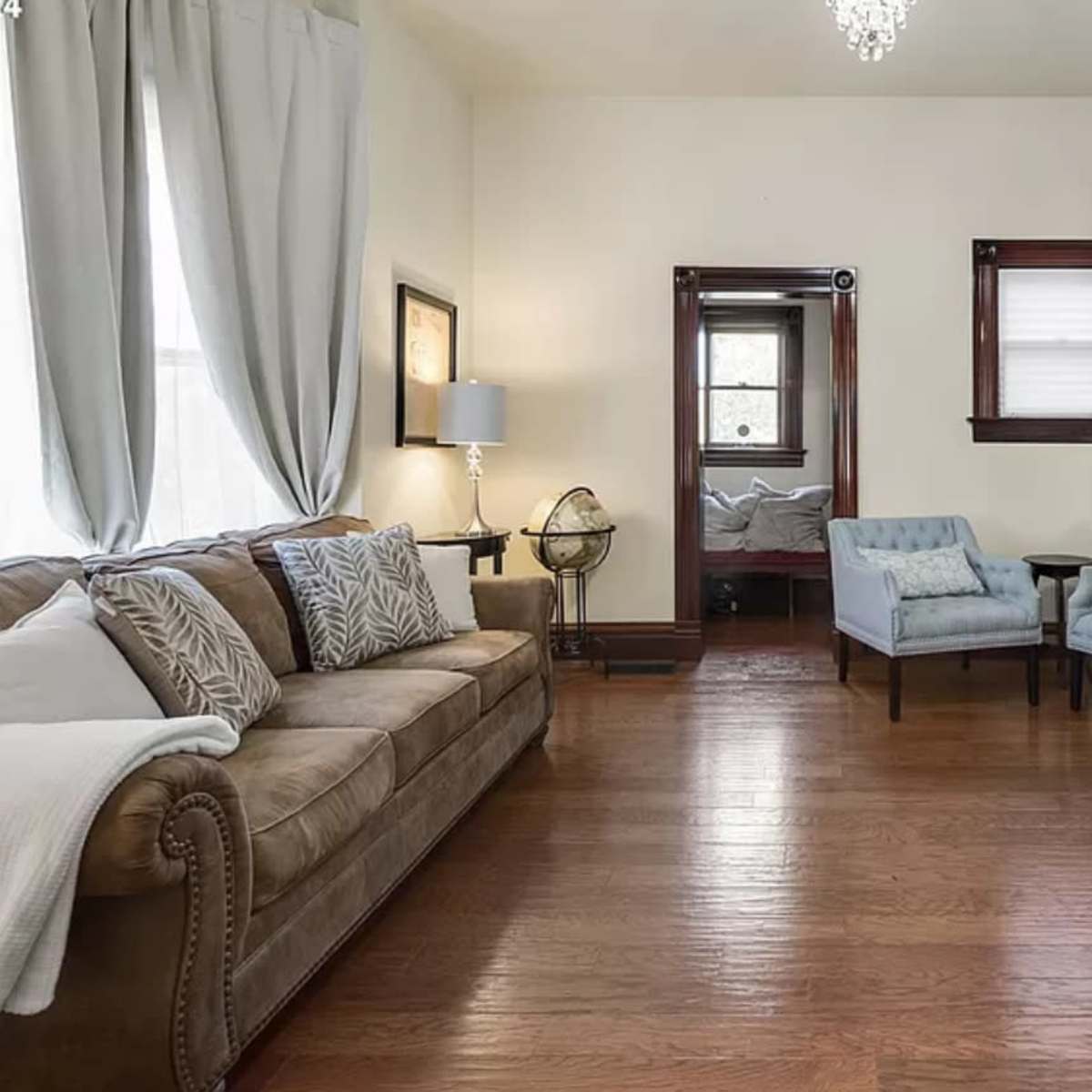 A living room with a brown sofa, two blue chairs, and wooden flooring, along with a small round table and a doorway leading to another room.