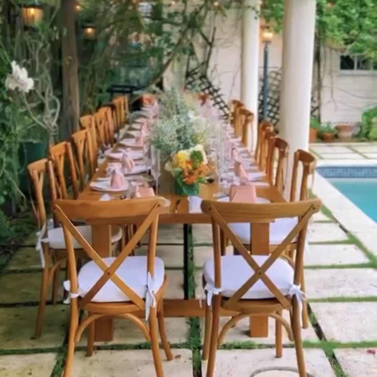 A long wooden dining table is set with place settings and flowers under a pergola, surrounded by greenery and near a pool.