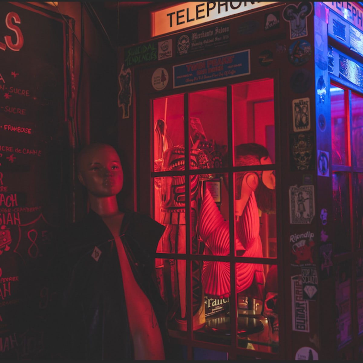 A brightly lit vintage telephone booth adorned with stickers stands next to a mannequin in a dimly lit space.