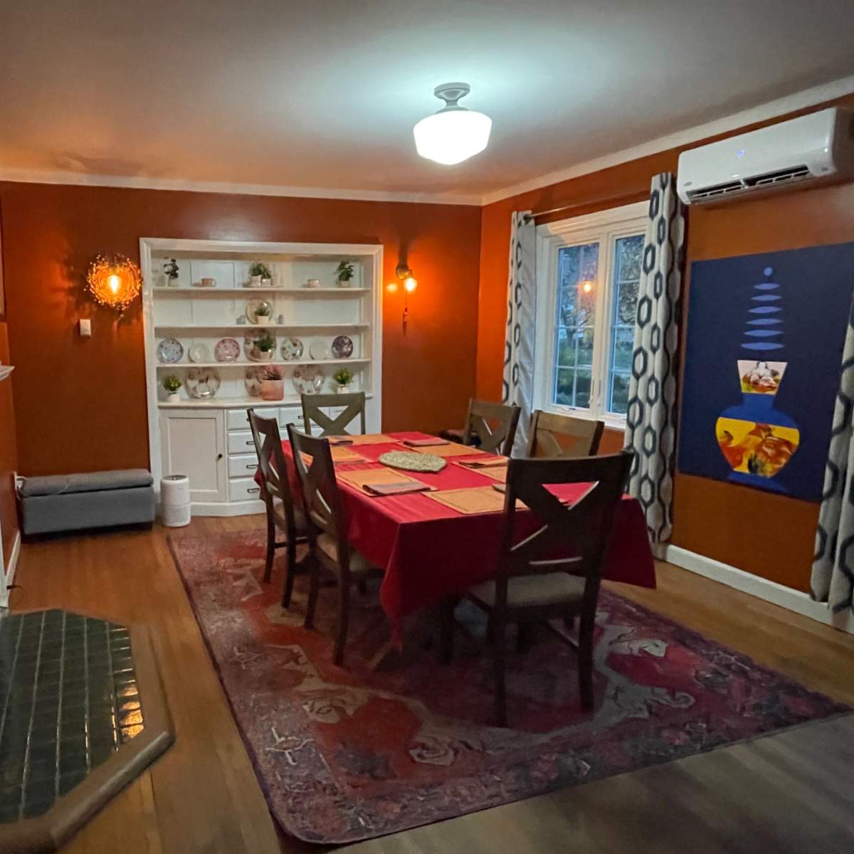 A dining room features a red tablecloth over a large table surrounded by chairs, with a fireplace and shelves displaying dishes along the walls.