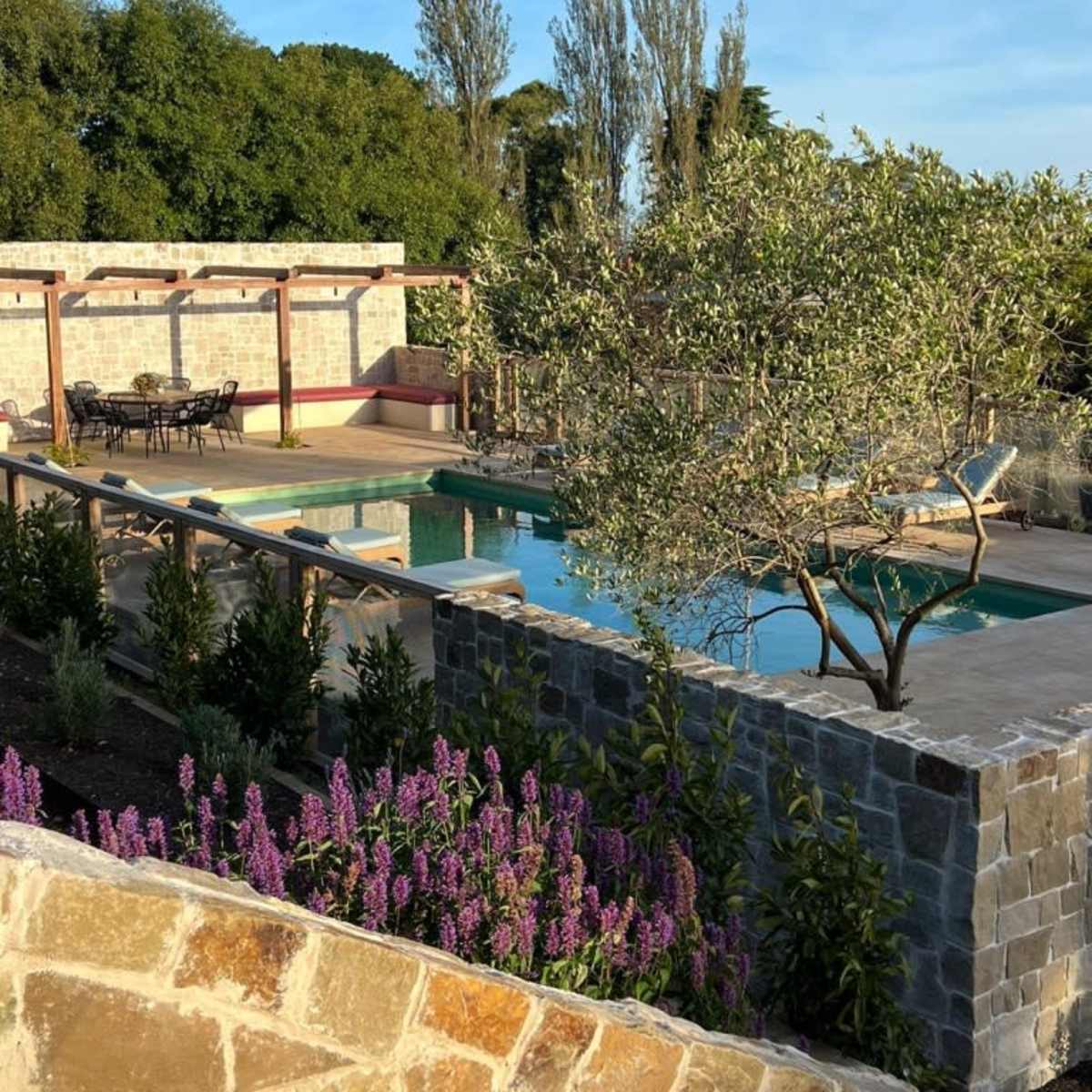 A landscaped area featuring a swimming pool surrounded by stone walls and vibrant purple flowers, with outdoor seating visible in the background.