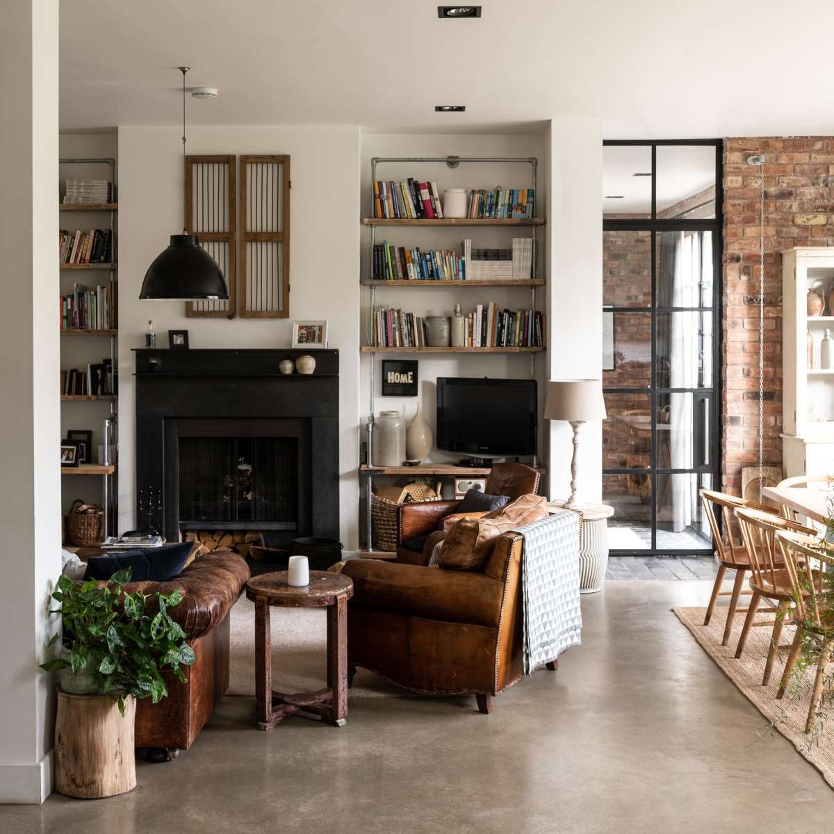 A modern living room featuring a fireplace, bookshelves filled with books, leather furniture, and a dining area with large windows and a wooden table.