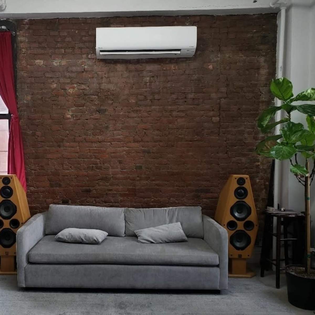 A gray sofa between two wooden speakers against a backdrop of a brick wall, with a window draped in red curtains and a potted plant to the right.