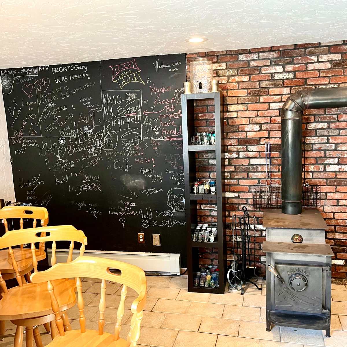 A room with a brick wall and a chalkboard covered in various drawings and writings, along with a cast iron stove and wooden chairs arranged nearby.