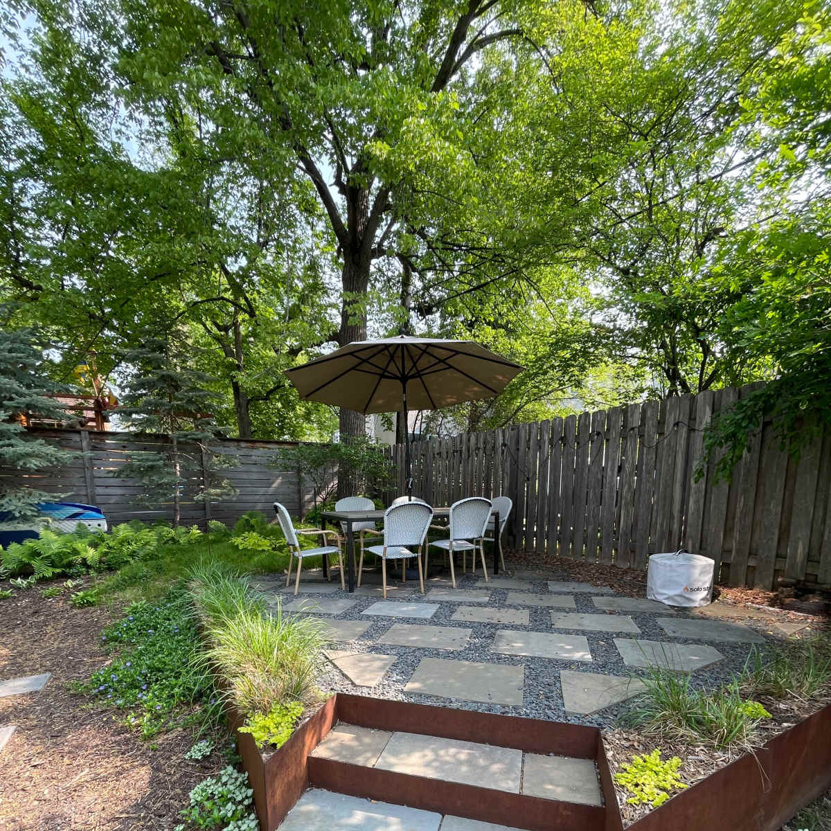 A patio area with a table and chairs is shaded by an umbrella, surrounded by greenery and bordered by a wooden fence.