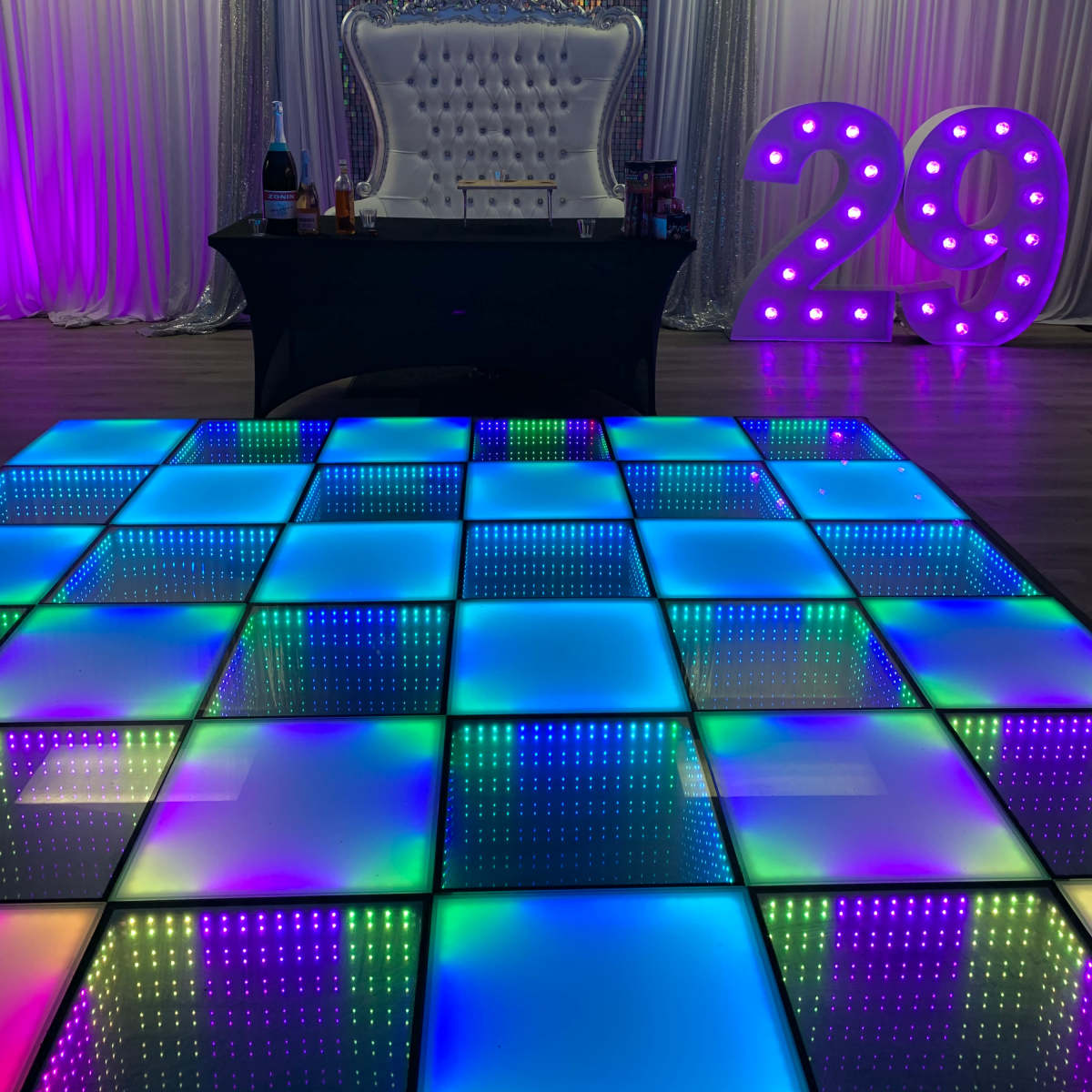 A decorated party space featuring a light-up dance floor, a bar setup, and a large illuminated "29" display against a backdrop of shimmering curtains.