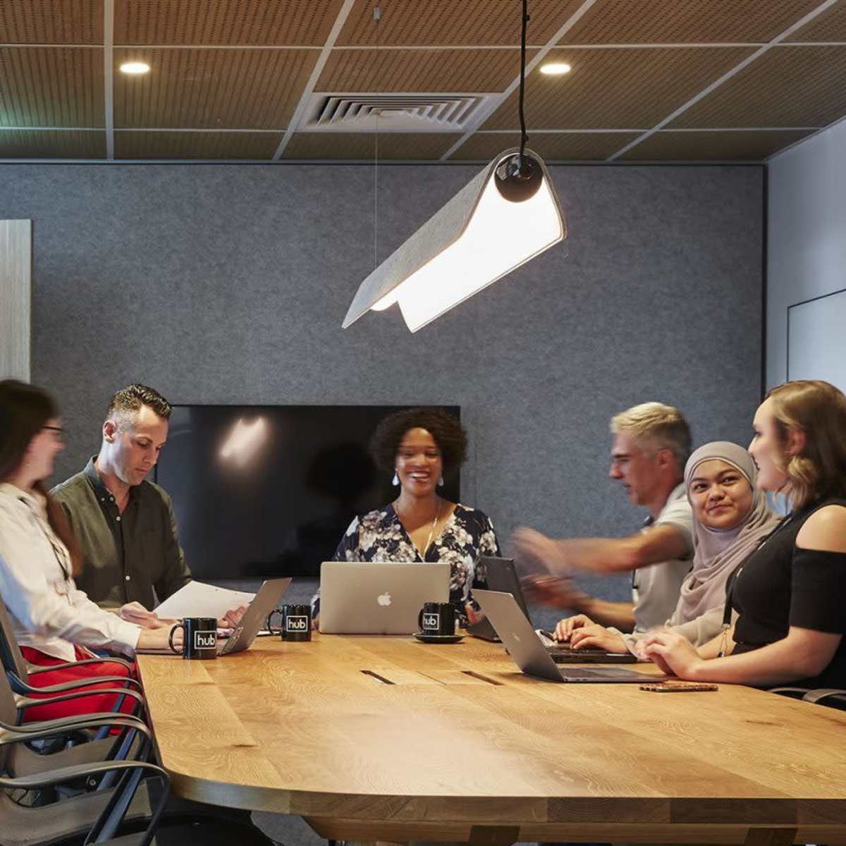 A group of seven people are sitting around a large wooden table in a modern meeting room, engaged in discussion while using laptops and papers.