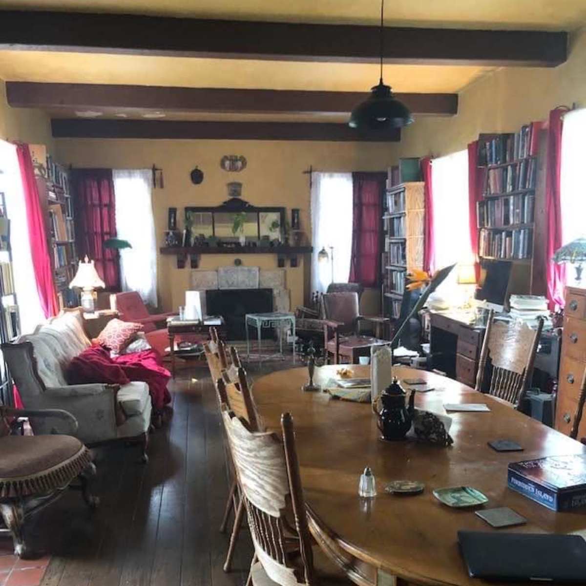 A spacious living room with a large wooden dining table, surrounded by chairs, bookshelves lining the walls, and a mix of furniture in a cozy arrangement.