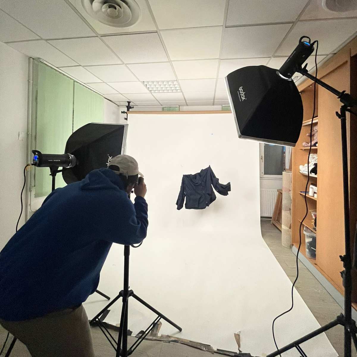 A person stands in front of a white backdrop, photographing a piece of clothing with studio lights set up on either side.