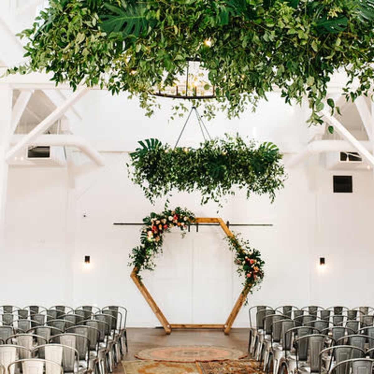 A wedding ceremony space with a geometric wooden arch adorned with greenery and flowers, surrounded by rows of metal chairs under hanging plants and lights.