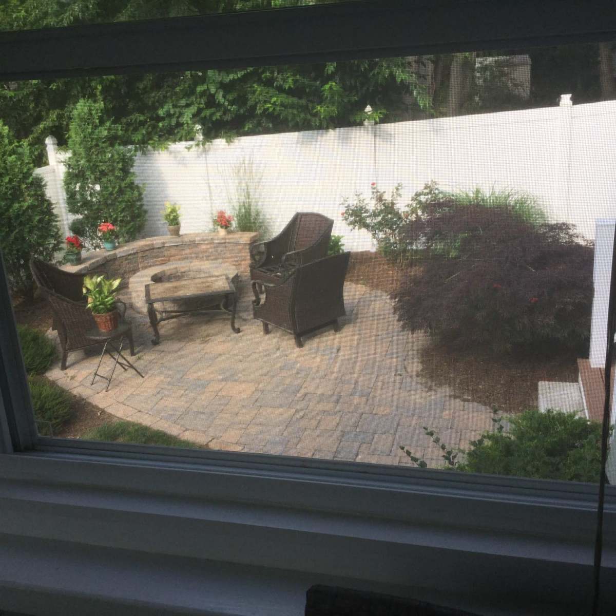 A stone patio features two wicker chairs and a small table surrounded by landscaped greenery and a white fence.