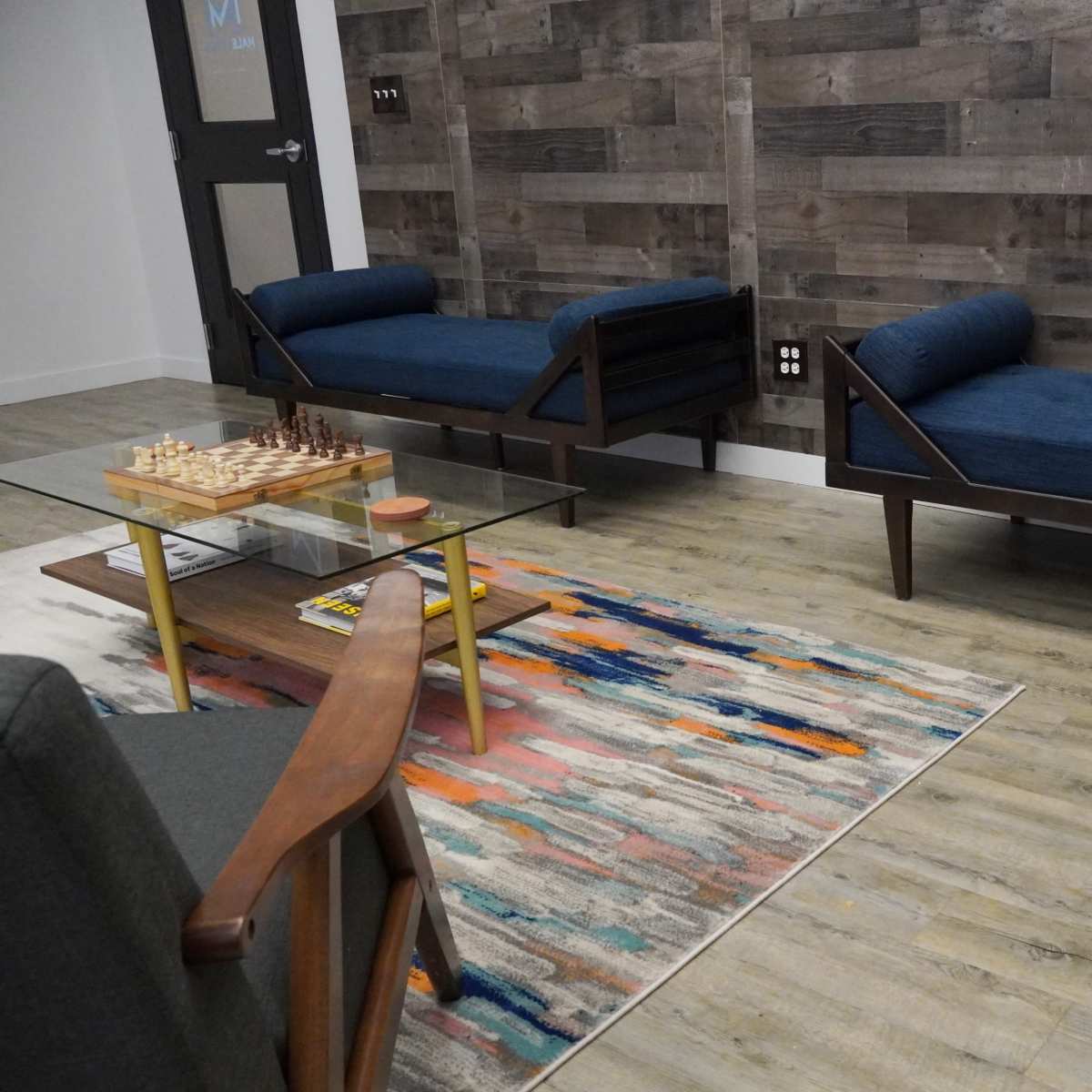 A modern lounge area with two blue upholstered couches, a glass coffee table on a colorful rug, and a chessboard set on the table.