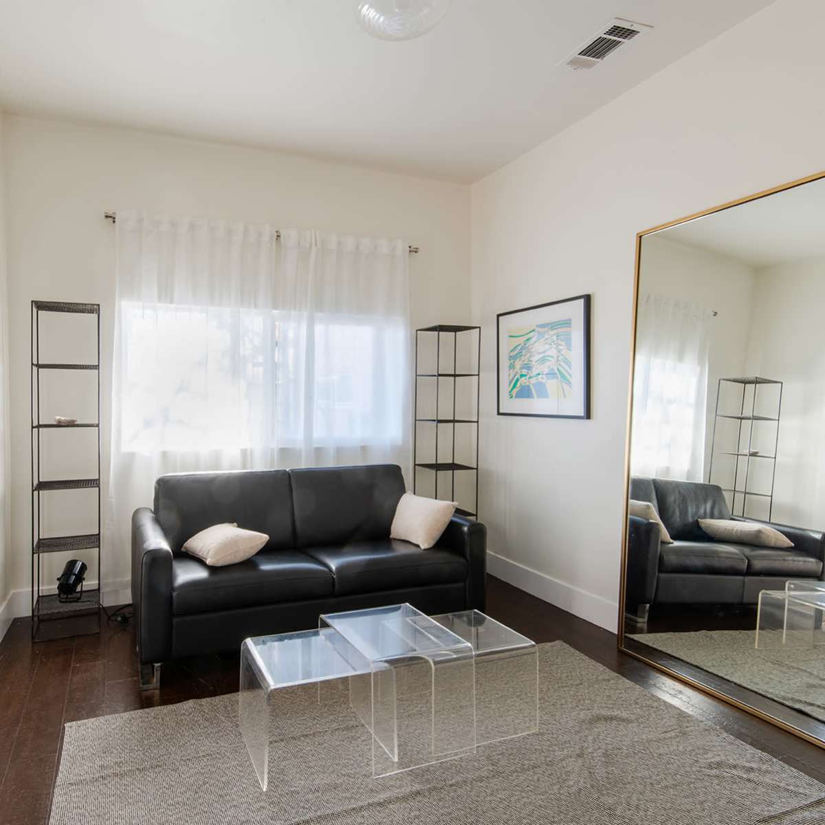 A living room with a black leather sofa, a large mirror, a glass coffee table, and a coat rack, illuminated by natural light from a window.