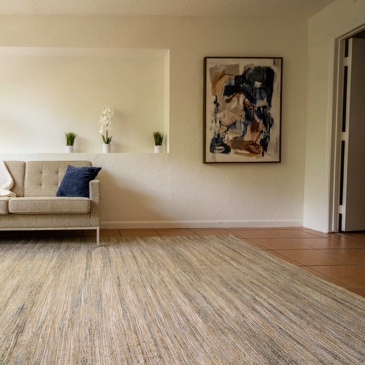 A simple living room featuring a light-colored sofa with blue pillows, a textured area rug, and a wall adorned with a framed artwork.
