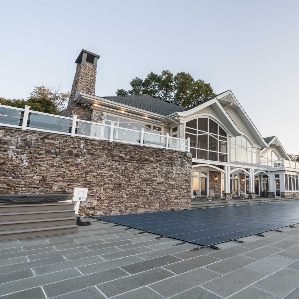 A large modern house with a stone exterior overlooks a pool area, featuring a covered pool cover and several lounge chairs arranged on a paved patio.