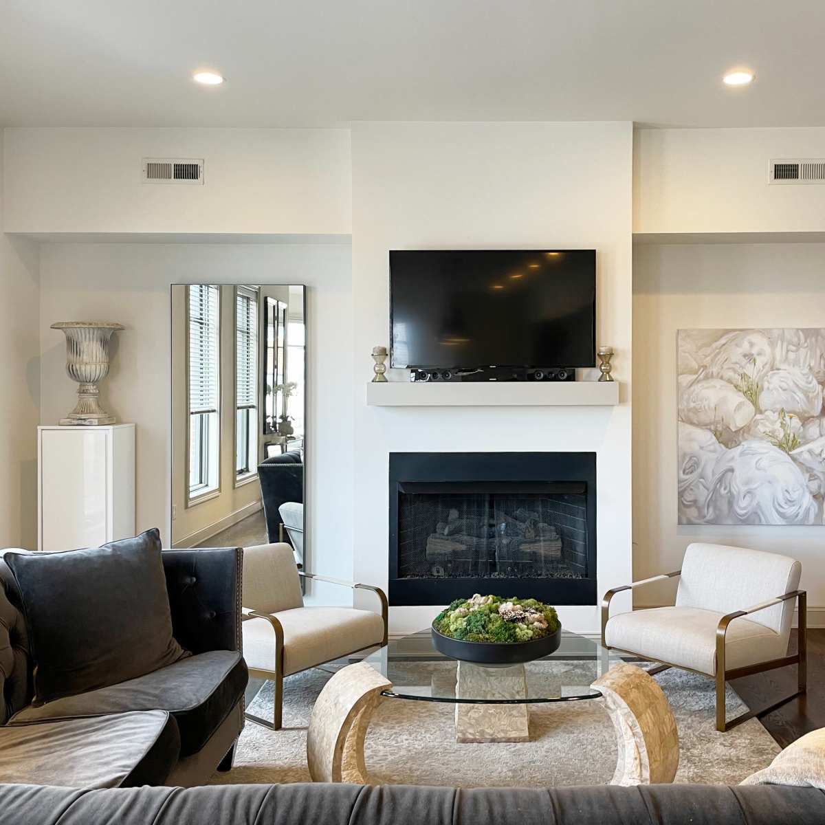 A modern living room features a fireplace, a wall-mounted TV, two armchairs, and a circular glass coffee table with a decorative centerpiece.