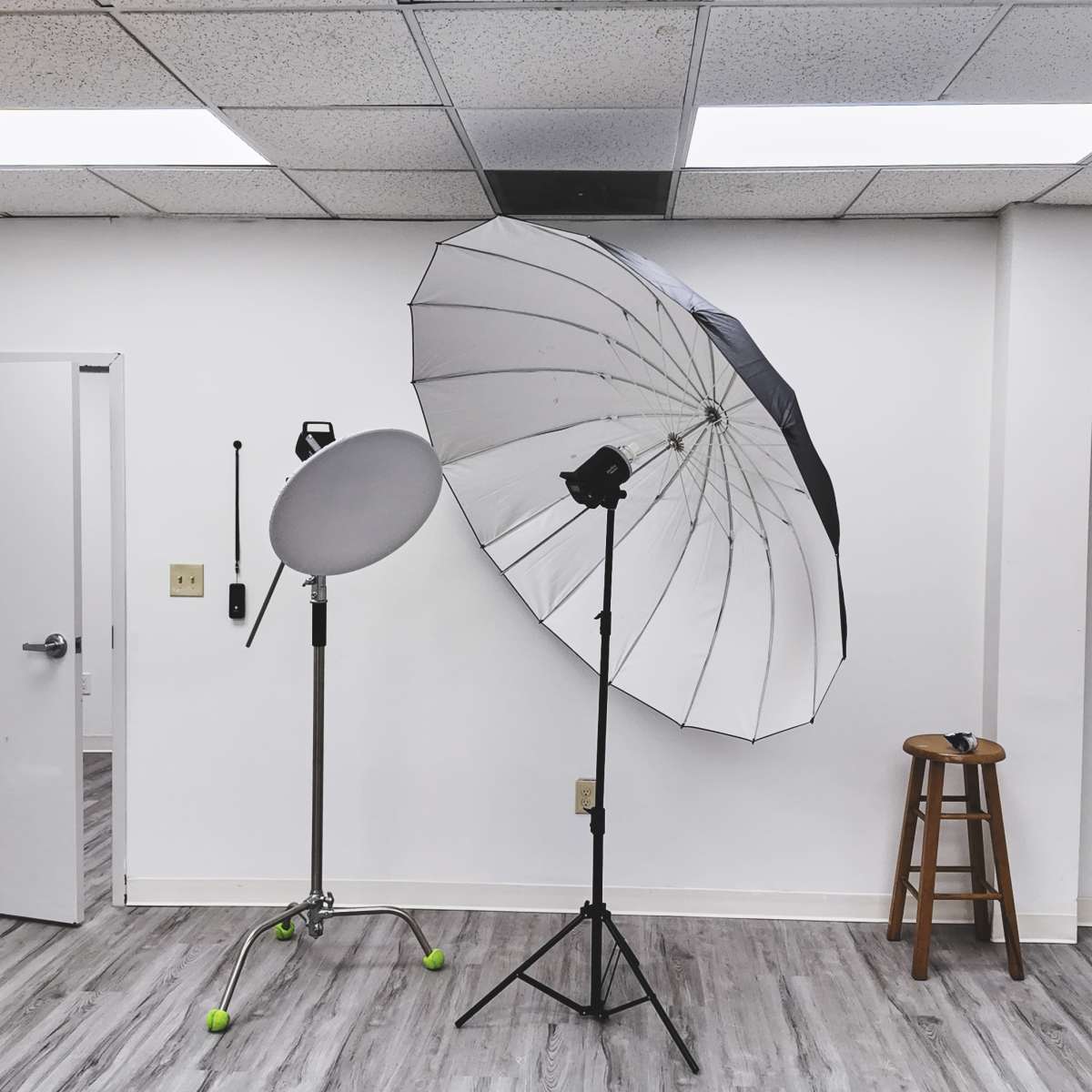 A photography studio with a large umbrella softbox, a reflector on a stand, a stool, and plain white walls.