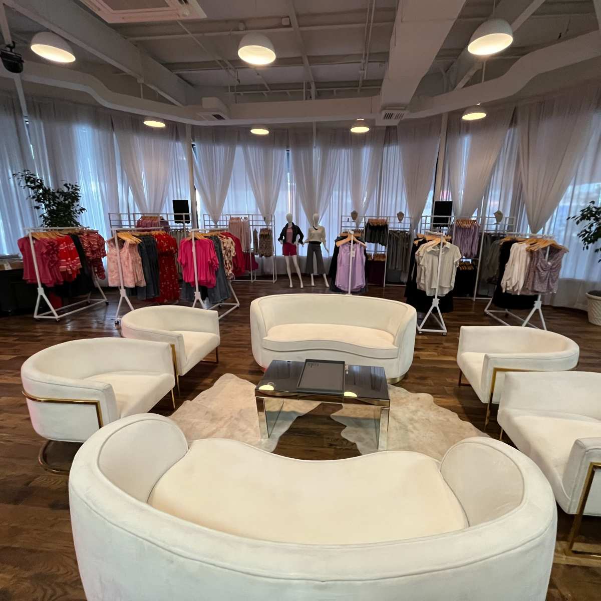 A modern clothing showroom features white seating arranged around a central table, with displays of colorful garments hanging on racks against the backdrop of large, airy windows.