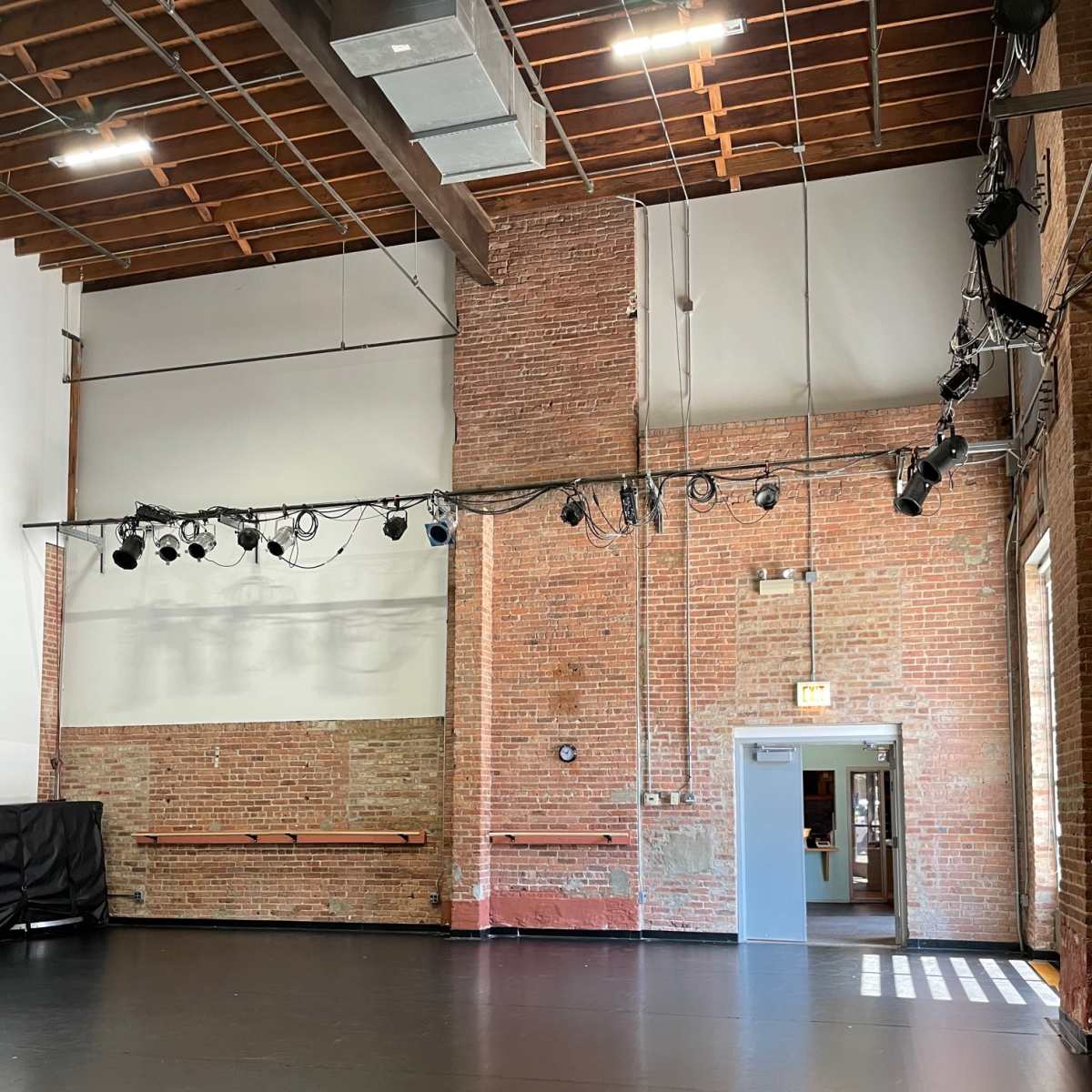 A spacious rehearsal studio with exposed brick walls, a polished black floor, and a row of overhead lights.
