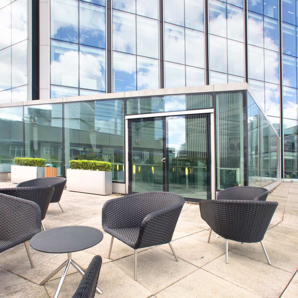 An outdoor seating area with dark wicker chairs and small tables in front of a modern glass building.