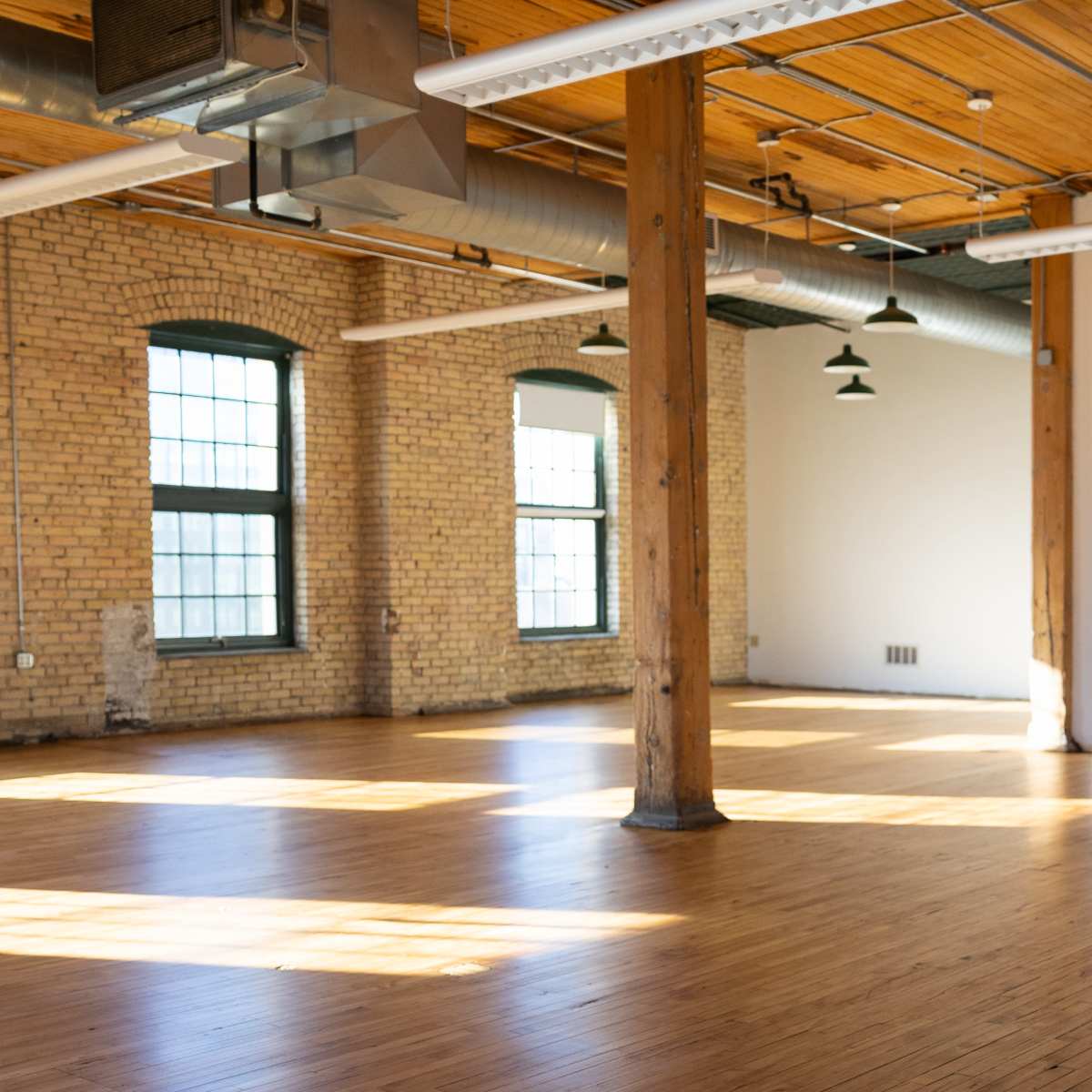 A spacious, empty room with wooden floors, high ceilings, and large windows allowing natural light to enter.