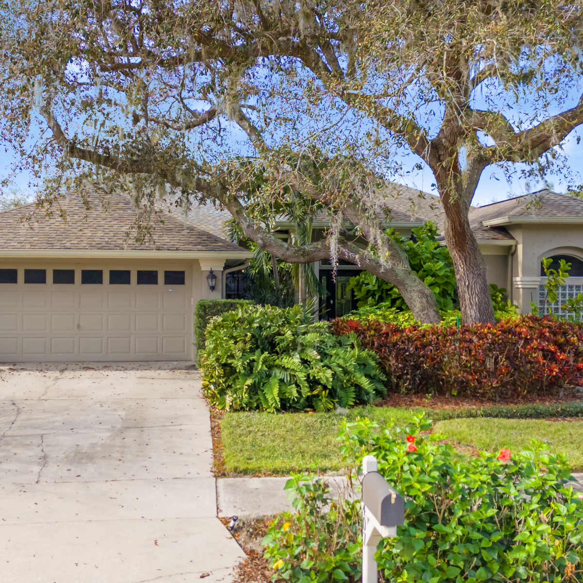 A house with a two-car garage is surrounded by landscaped greenery and a large tree in the front yard.