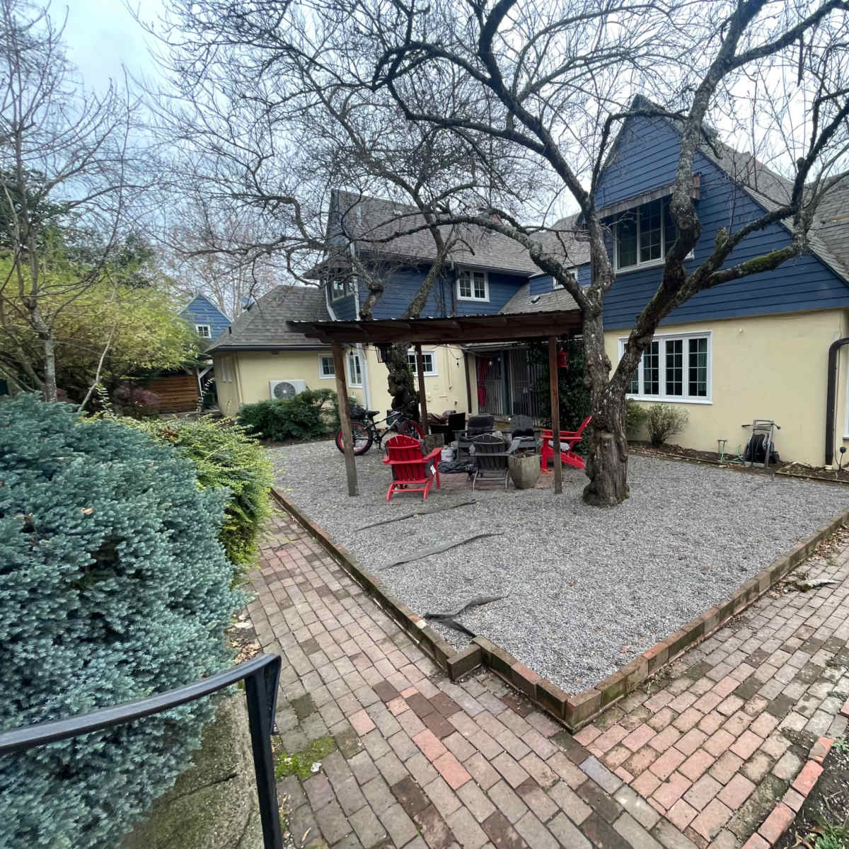 A backyard with a gravel area surrounded by trees, a patio with red chairs, and a house featuring a mix of blue and beige siding.