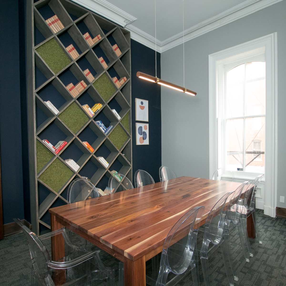 A modern meeting room featuring a large wooden table surrounded by transparent chairs, with a geometric bookshelf filled with books and decorative elements in the background.