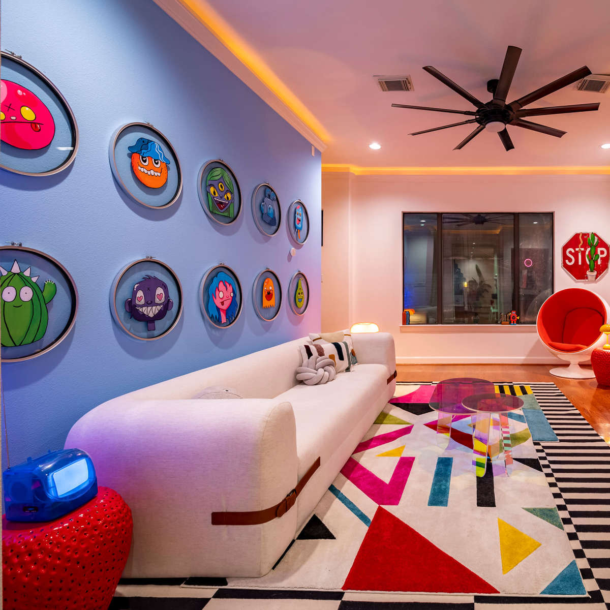 The room features a colorful modern design with a white sofa, a bold area rug, framed artwork on the walls, and a large window.