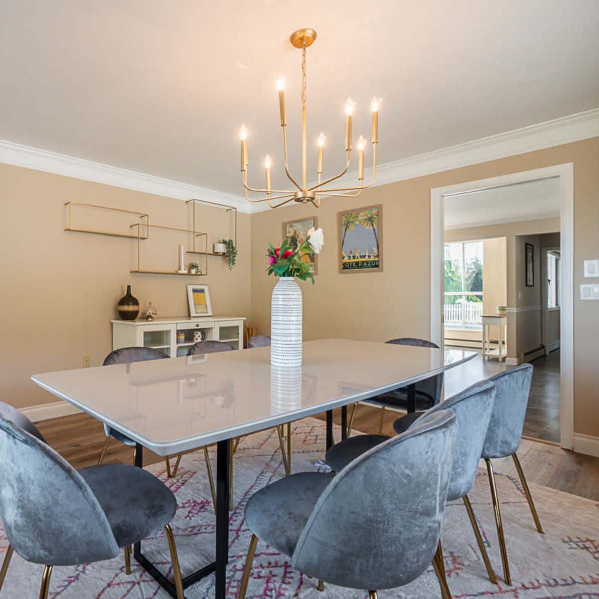 A modern dining room features a rectangular table surrounded by six upholstered chairs, illuminated by a chandelier, with a decorative vase and artwork on the walls.