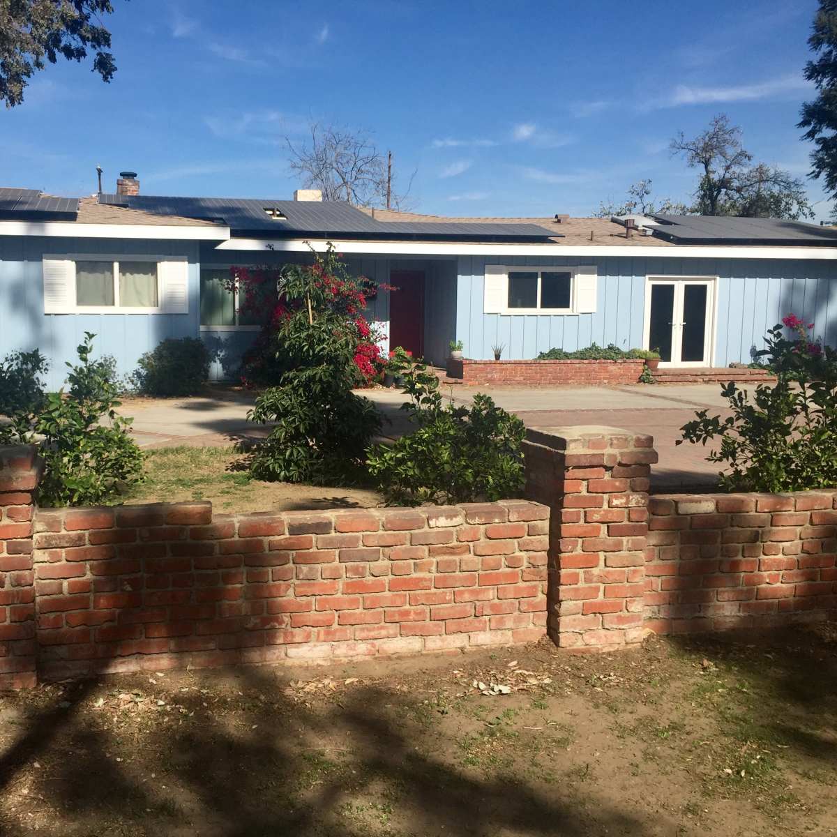 A single-story blue house with a red door is surrounded by a brick wall and landscaped with shrubs and trees.