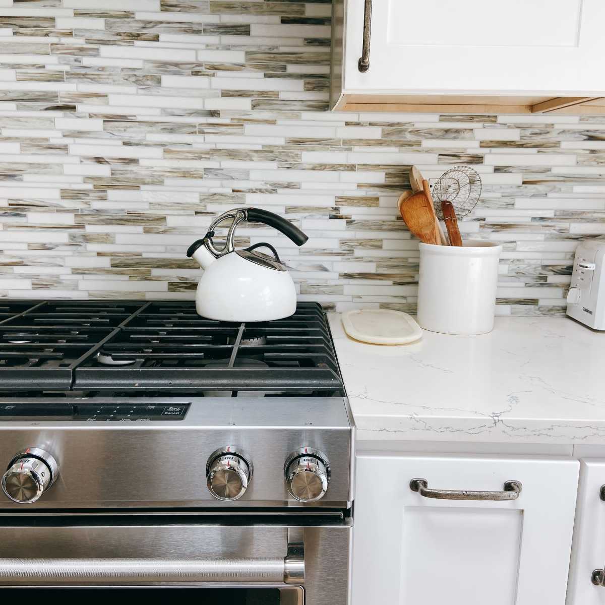 A stainless steel gas stove with a black-handled kettle rests on a marble countertop, set against a textured mosaic tile backsplash.