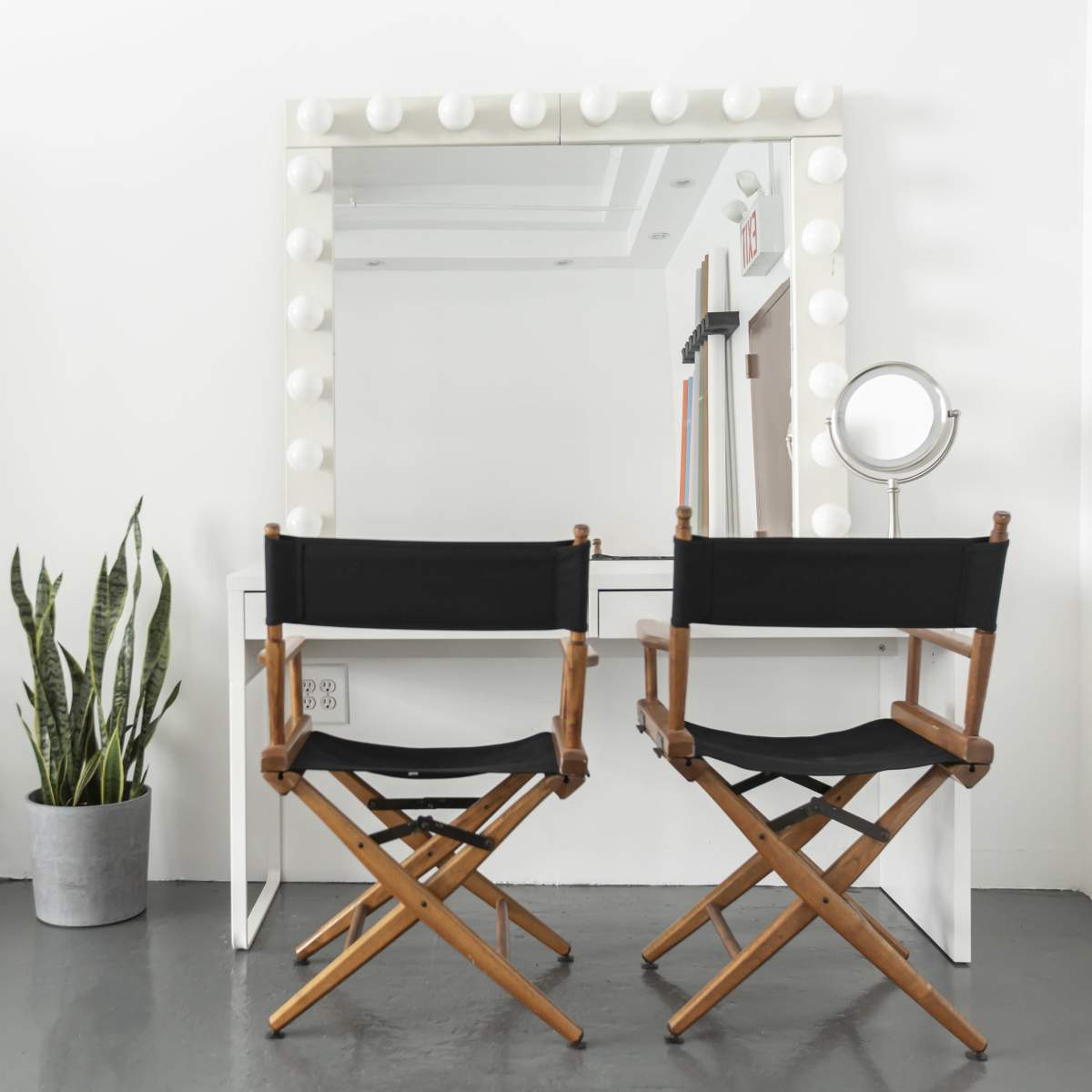 A minimalist dressing area with two wooden director's chairs facing a large illuminated mirror, a small round mirror, a potted plant, and a fan nearby.