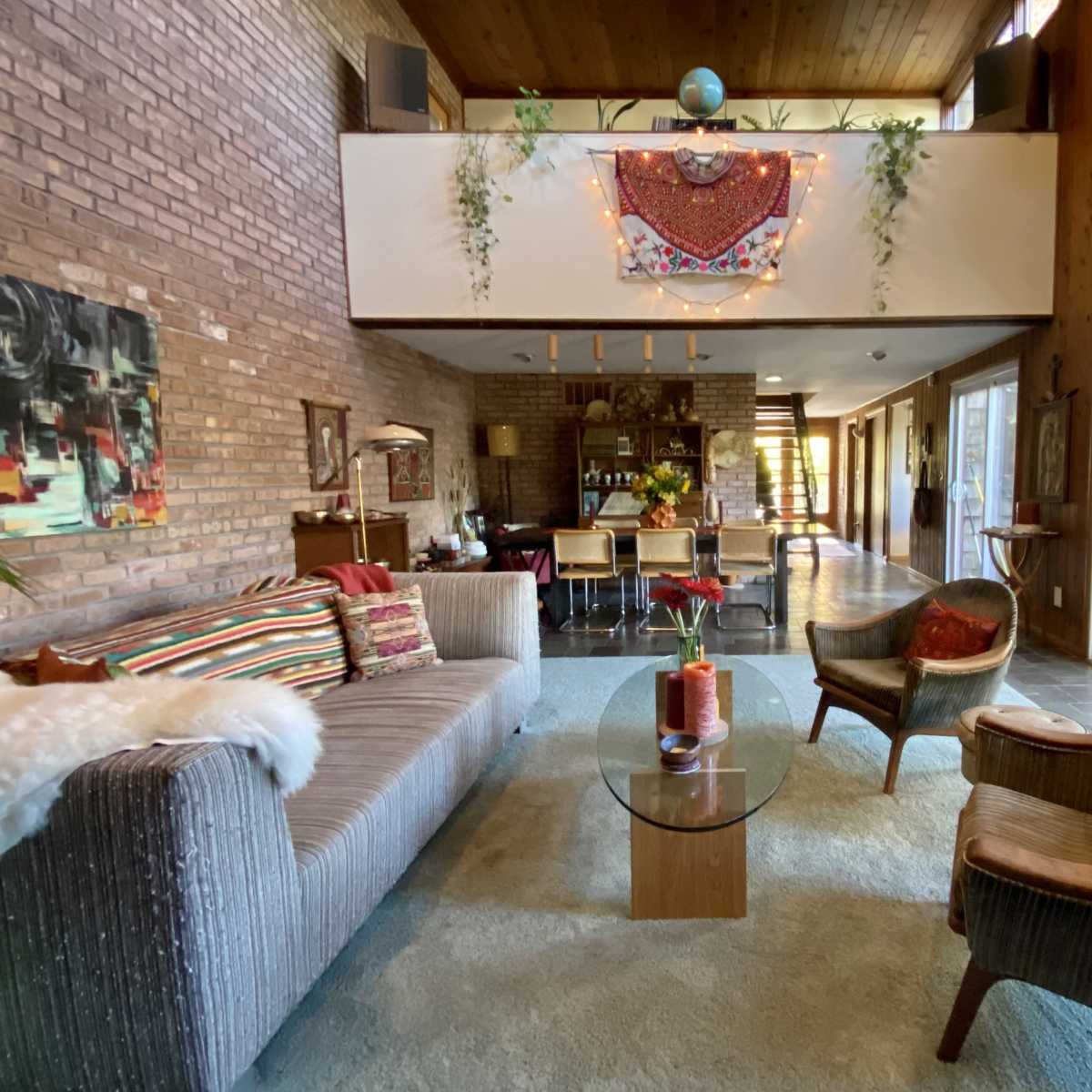 A modern living room with a sofa, armchairs, a glass coffee table, and large windows that open to a hallway and kitchen area.