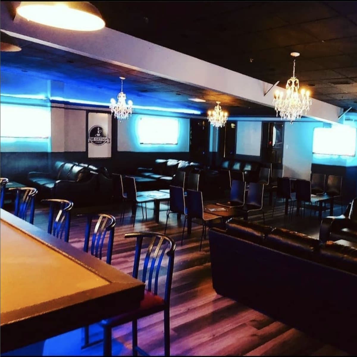 A spacious lounge area with black leather seating, small tables, and chandeliers, illuminated by blue lighting.