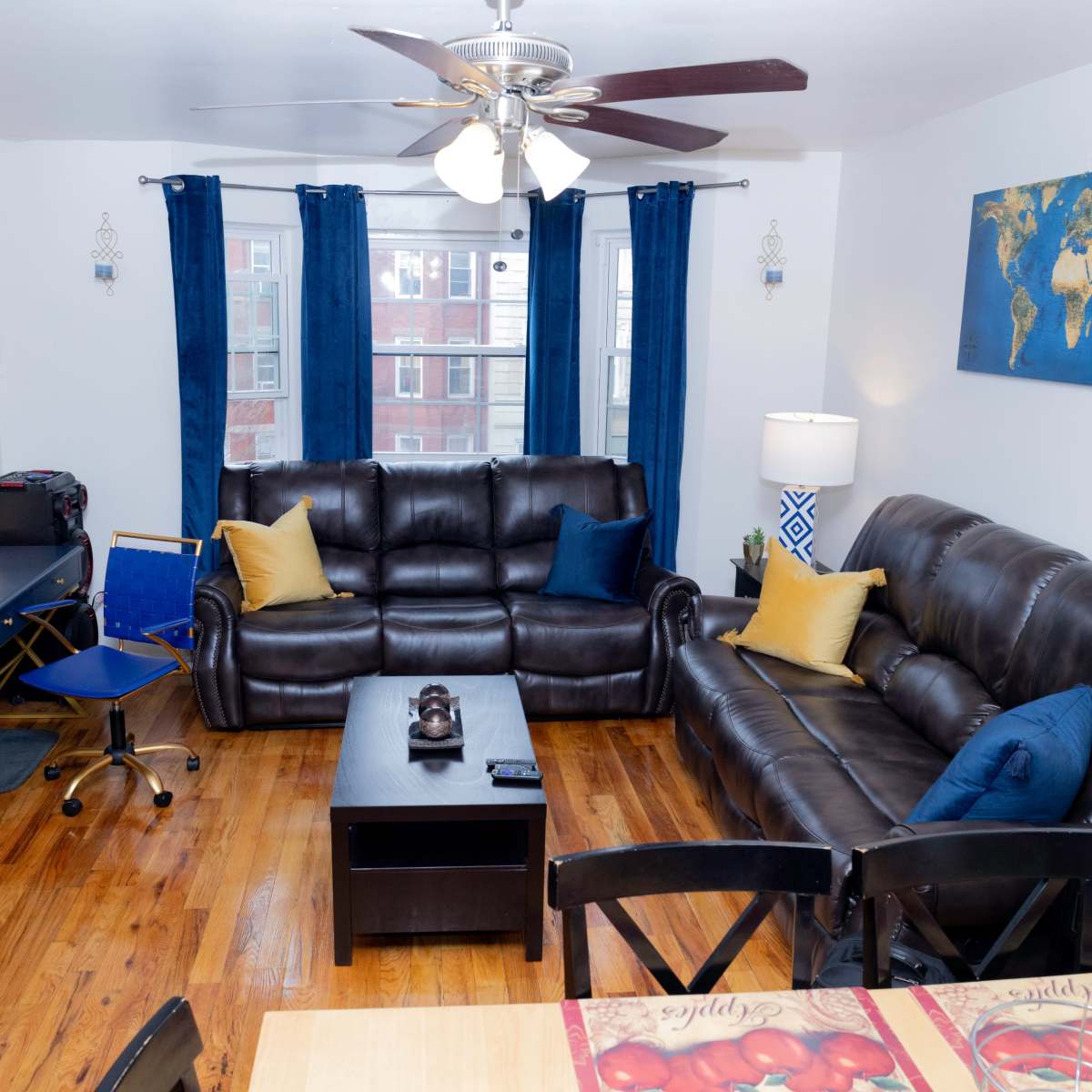 A living room features two dark leather sofas, a coffee table, a TV stand, and a desk, with blue curtains framing the windows and a world map on the wall.