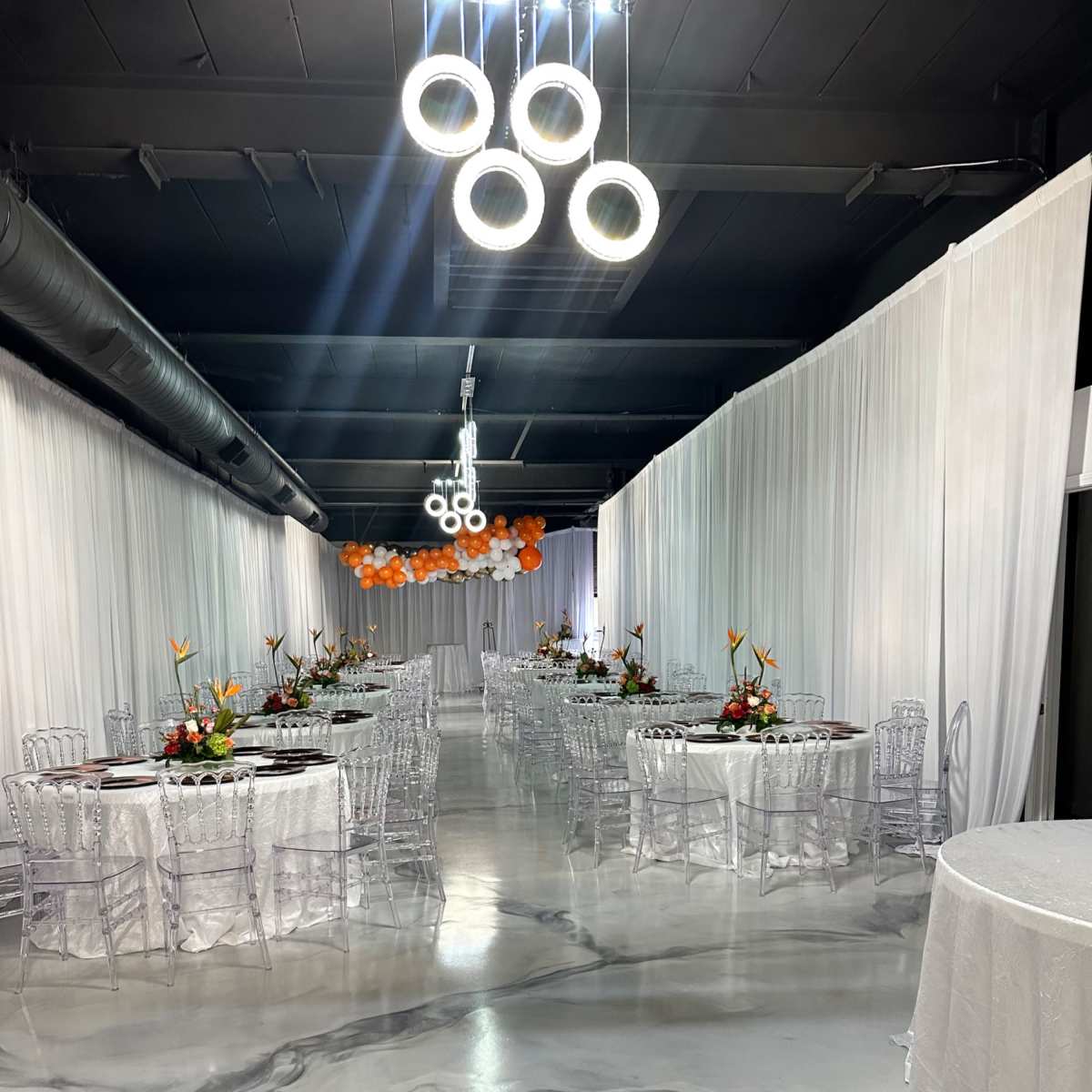 A spacious event hall with white draped walls, circular chandeliers, and decorated tables arranged in two rows.