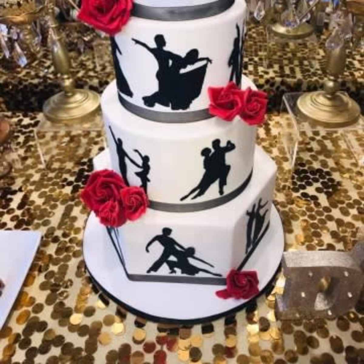 A three-tiered cake decorated with black silhouettes of dancers and red roses, set against a table with a sparkling gold sequin tablecloth.