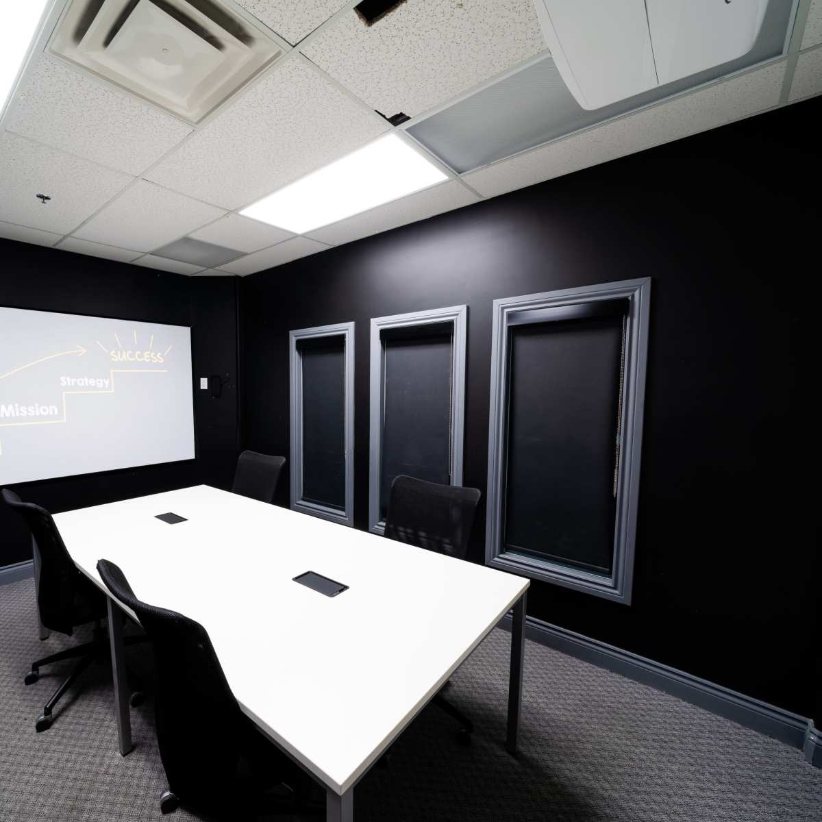 A modern conference room with a large white table and black chairs, featuring a black wall and a presentation screen displaying the words "Success," "Strategy," "Mission," and "Vision."