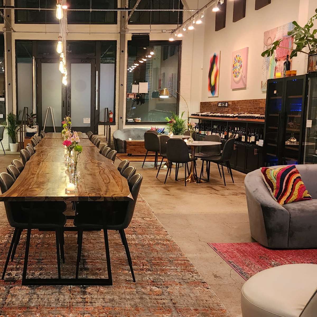 A spacious interior of a modern venue with a long wooden dining table surrounded by chairs, a bar area with high stools, and various decor elements including plants and artwork.