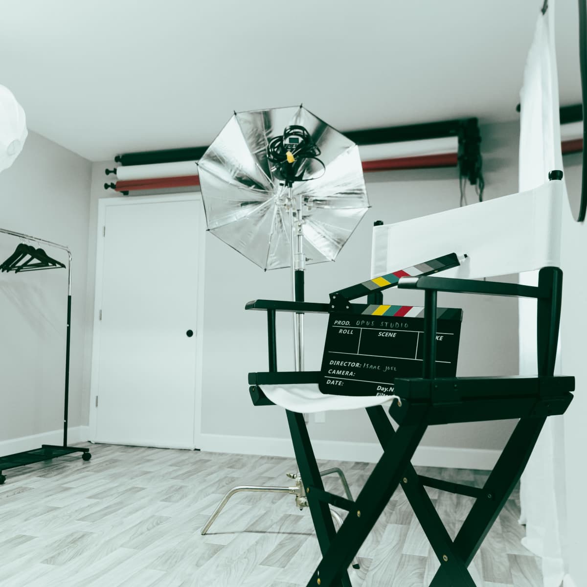 A film studio setup features a director's chair, a clapperboard, lighting equipment, and a clothing rack against a plain wall.