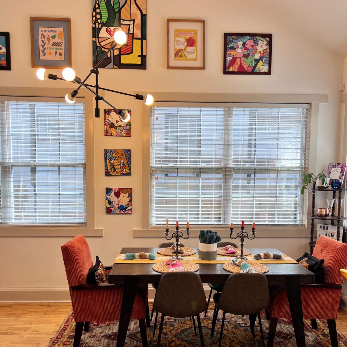 A dining area features a table set with decorative plates and candles, surrounded by colorful chairs and adorned with framed artwork on the walls.