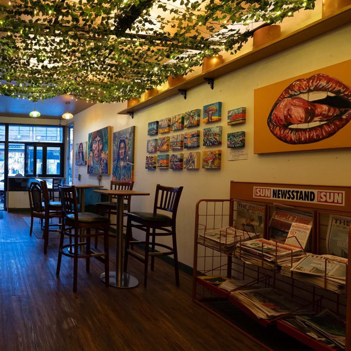 The interior of a cafe features wooden tables and chairs, vibrant artwork on the walls, and a display of newspapers near the entrance.