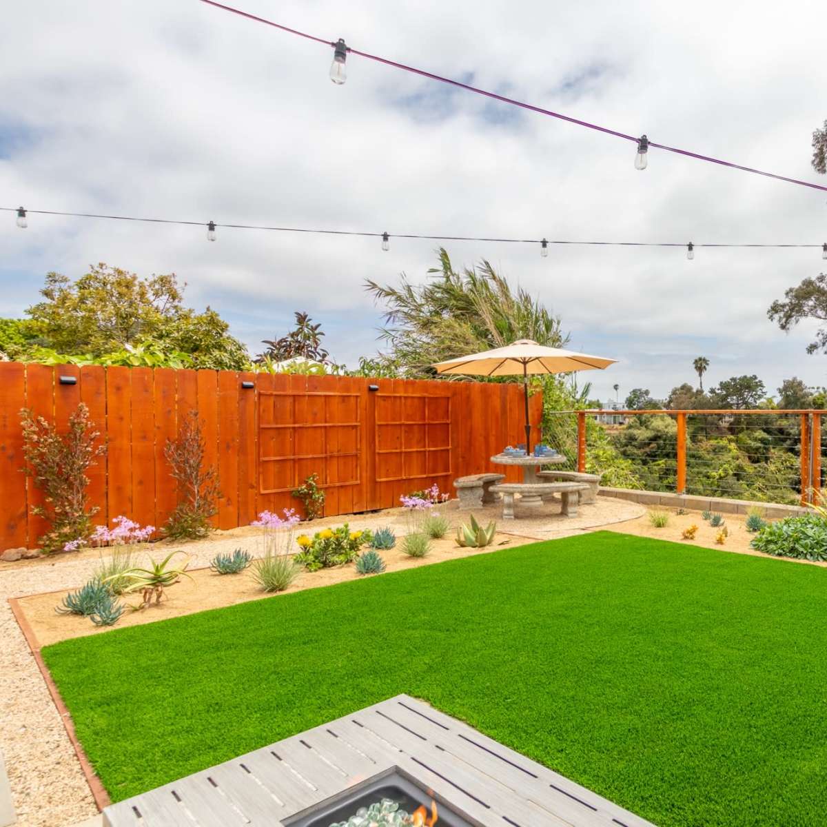 A landscaped backyard with a well-maintained lawn, surrounded by a wooden fence and a glass railing, featuring a table under an umbrella and decorative plants.
