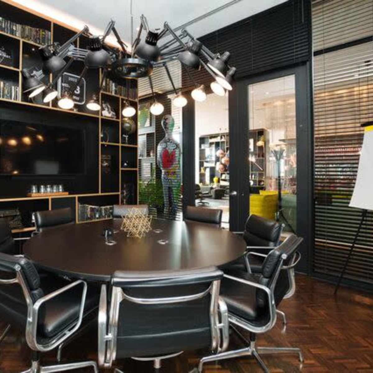 A modern conference room features a round table surrounded by sleek black chairs, a large TV, and wall-mounted bookshelves filled with various items.